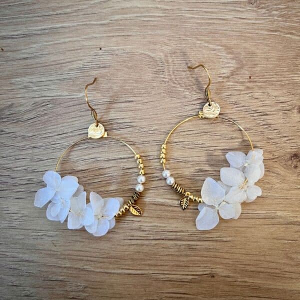 Les boucles Lucie dévoilent de véritables pétales d’hortensias couleur blanche, stabilisés et suspendus à des créoles dorées avec de jolies petites breloques.