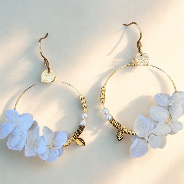 Les boucles Lucie dévoilent de véritables pétales d’hortensias couleur blanche, stabilisés et suspendus à des créoles dorées avec de jolies petites breloques.