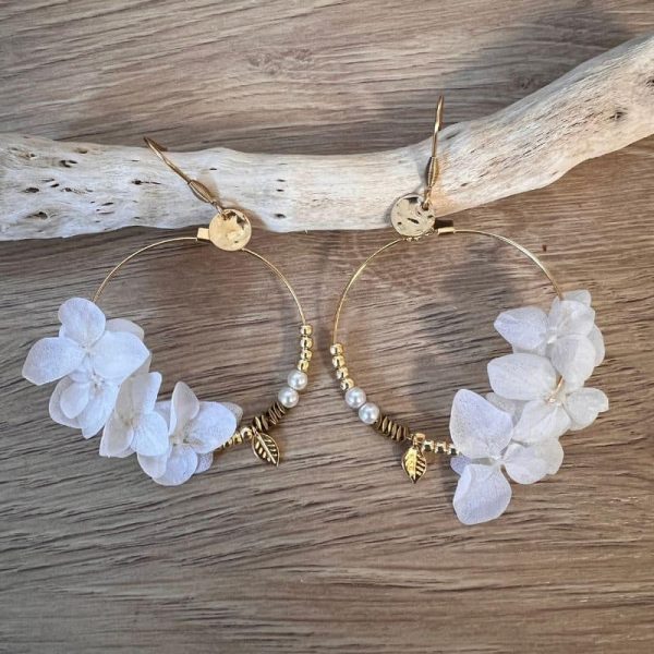 Les boucles Lucie dévoilent de véritables pétales d’hortensias couleur blanche, stabilisés et suspendus à des créoles dorées avec de jolies petites breloques.